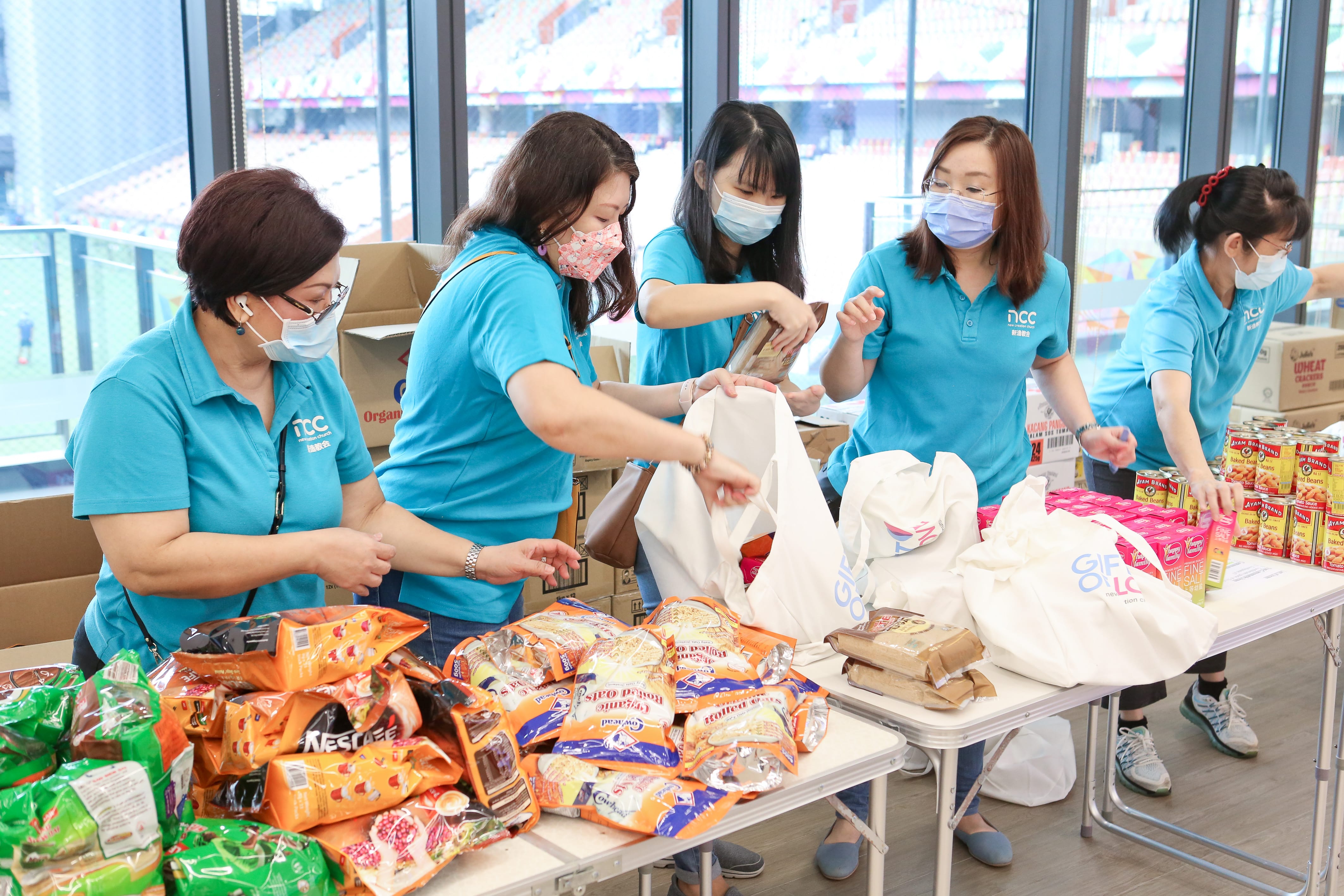 People in blue shirts pack food items into bags on tables filled with snacks and boxes.